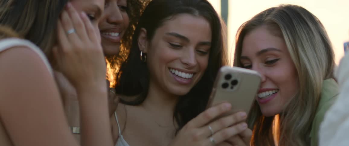 Three women laugh and clap while a fourth woman smiles and looks at a cell phone while standing outdoors