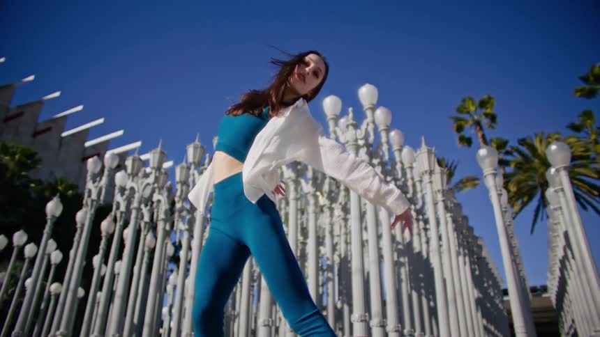 Young woman wearing blue clothes and white shirt energetically dancing in front of streetlamp on a park in the city