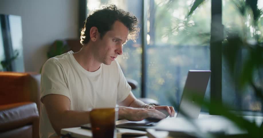 Man with white t shrt sits at desk using smartphone and laptop next to brown couch in living room at home