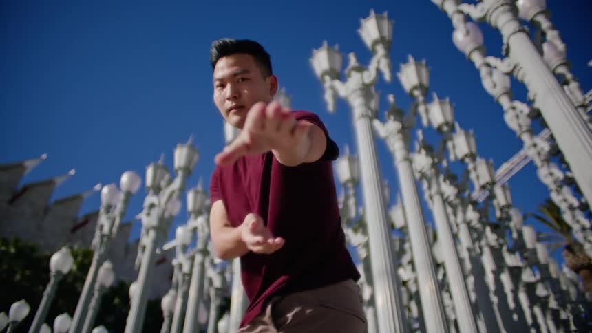 An asian male looks directly at the camera as he performs a dance outdoors