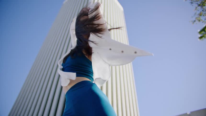 Woman wearing a blue outfit and white shirt dances in front of a building as the wind blows her hair