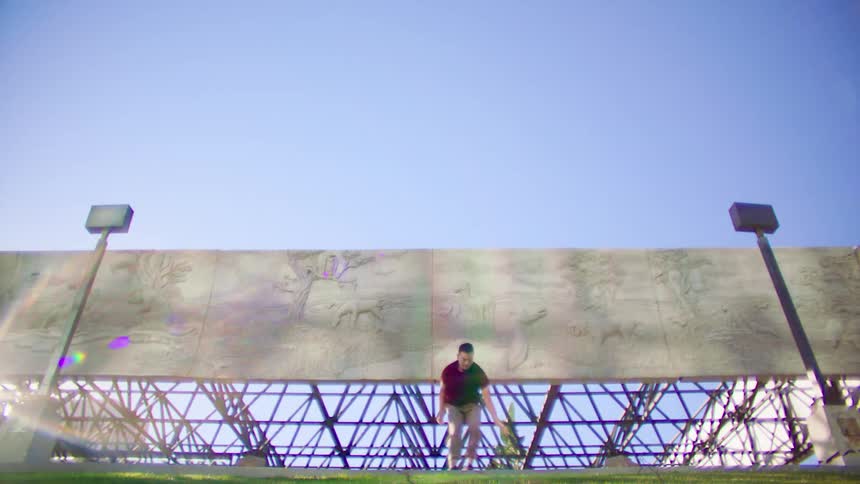 Man in a red short sleeved shirt dances in front of cement murals under a blue sky
