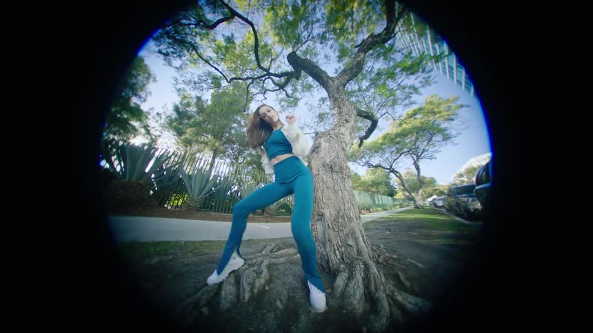 Young woman energetically dancing in front of a tree in a park at morning through a fisheye lens