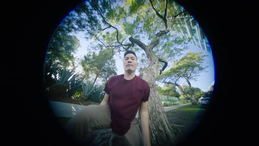 Young asian man energetically dancing in front of a tree in a park at morning seen through a fisheye lens