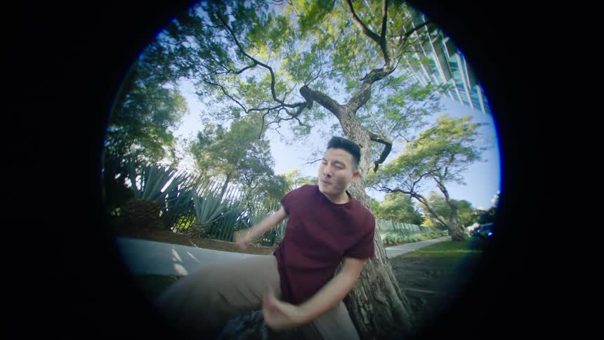 Male dancer dances on fish eye camera near tree growing near sidewalk