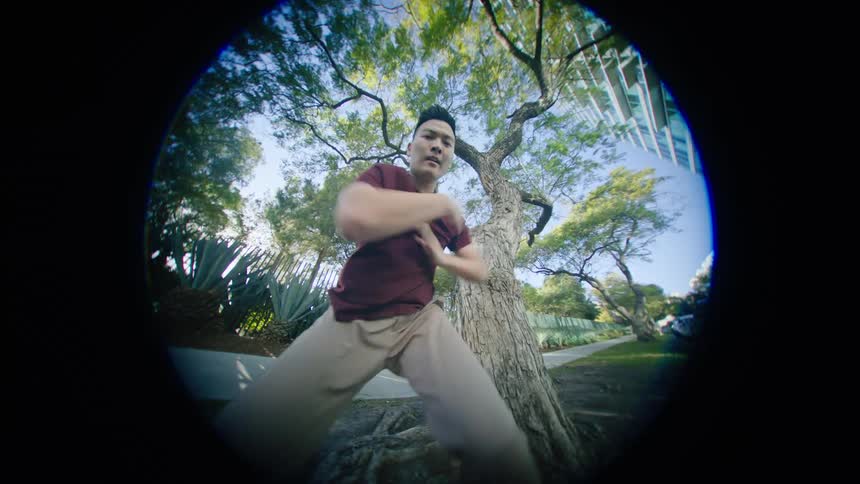 Young asian man energetically dancing in front of a tree in a park at morning through a fish eye lens