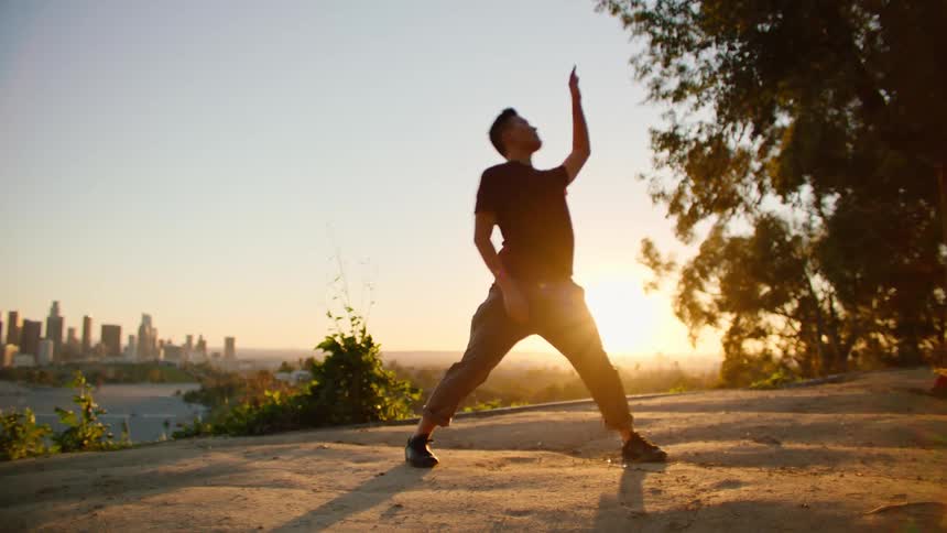 Dancing man wearing black t shirt and brown trousers is silhouetted against the city skyline and sunset