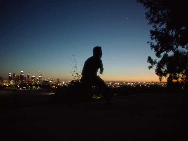 The silhouette of a man turning and leaping on a ridge overlooking a city at dusk