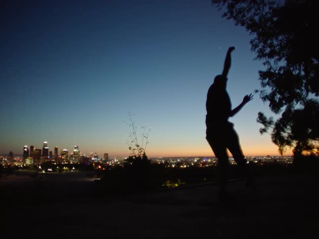 Man spins around while he dances outdoors as a city sits in the distance at night