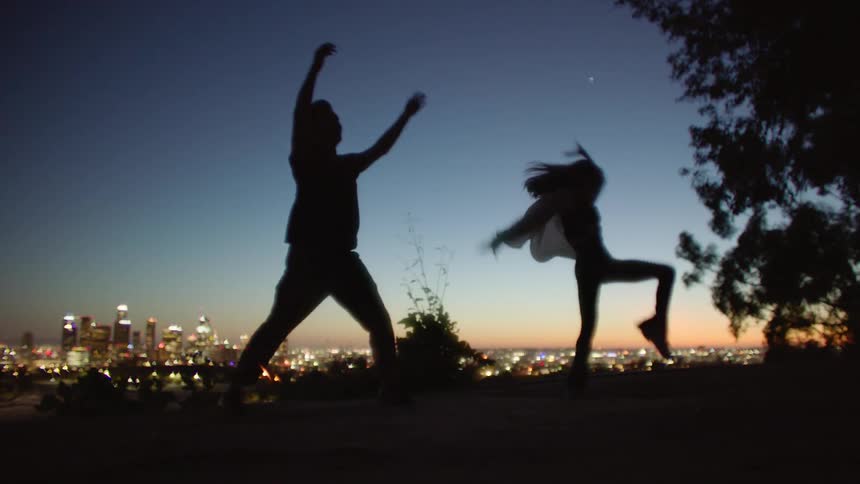 Man and woman perform dance moves outdoors at night near a tree with city buildings in the distance