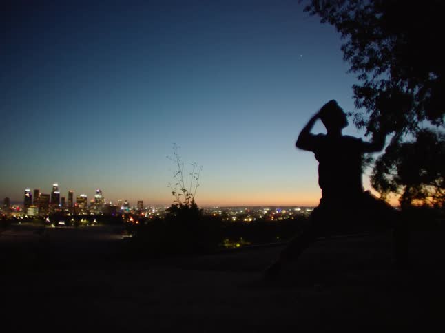Man jumps and spins as he dances outdoors at night with the city in the distance