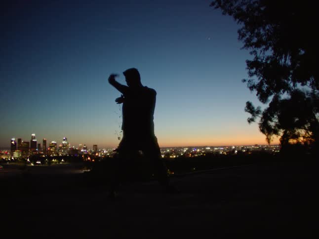 A man stretches and does a hip hop style dance while in a park with views of downtown