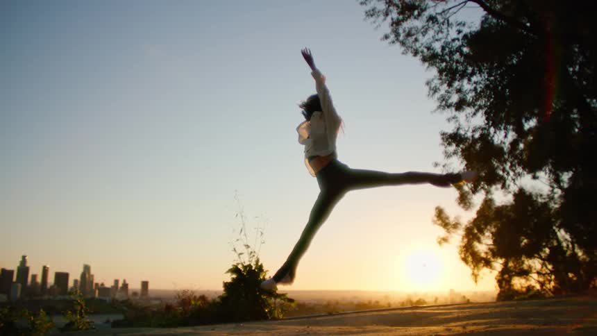Female dancer with white shirt and teal top and pants dances against city skyline at sunset