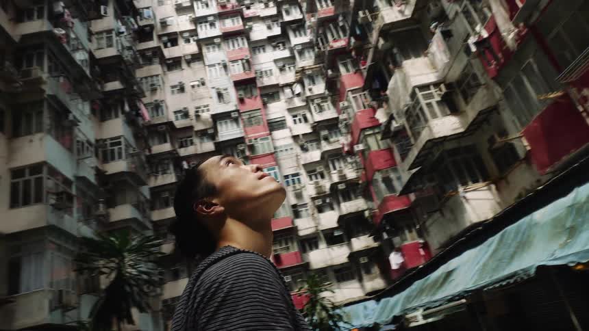 A young asian man stands on the ground and looks up at an apartment building and turns in a circle