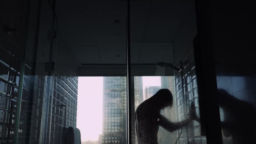 Young tired depressed man takes a shower in front of a window showing the city skyline