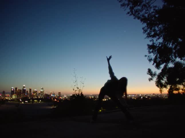 Woman stretches her arms out as she dances outdoors in the dark as city buildings sit in the distance