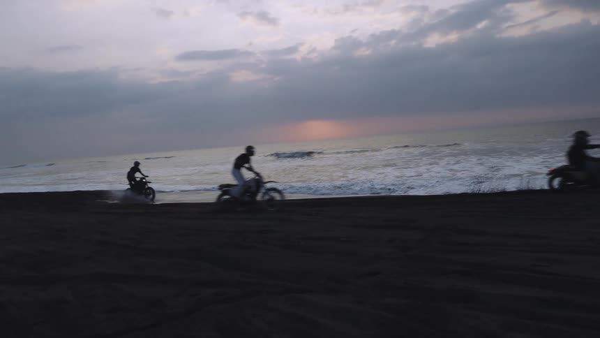 Three people ride motorcycles on the beach near the ocean at dusk