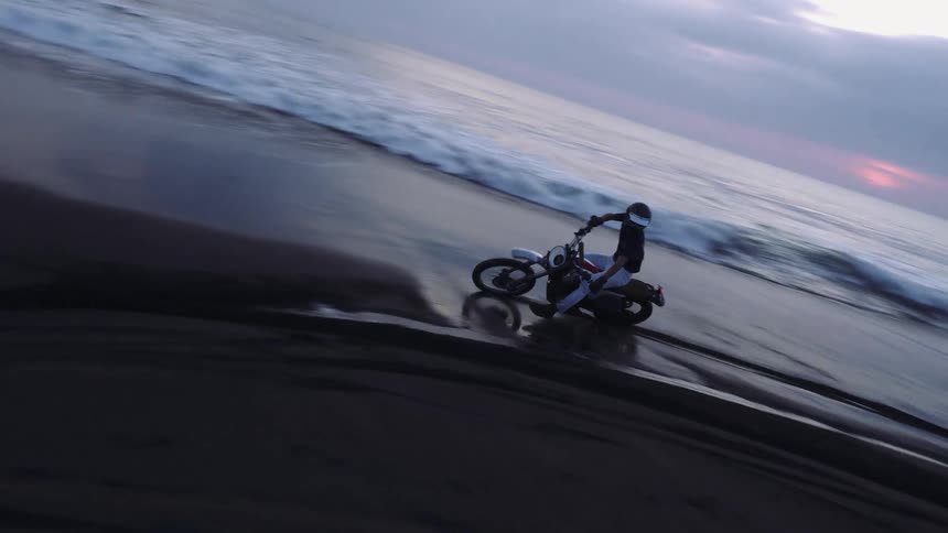 Male biker with helmet black shirt and jeans rides motorcycle on the beach next to sea at dusk