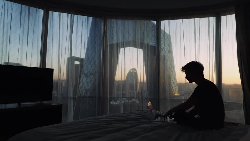 Man sits on a bed with a laptop as a television sits across the room and sheer curtains hang in the windows