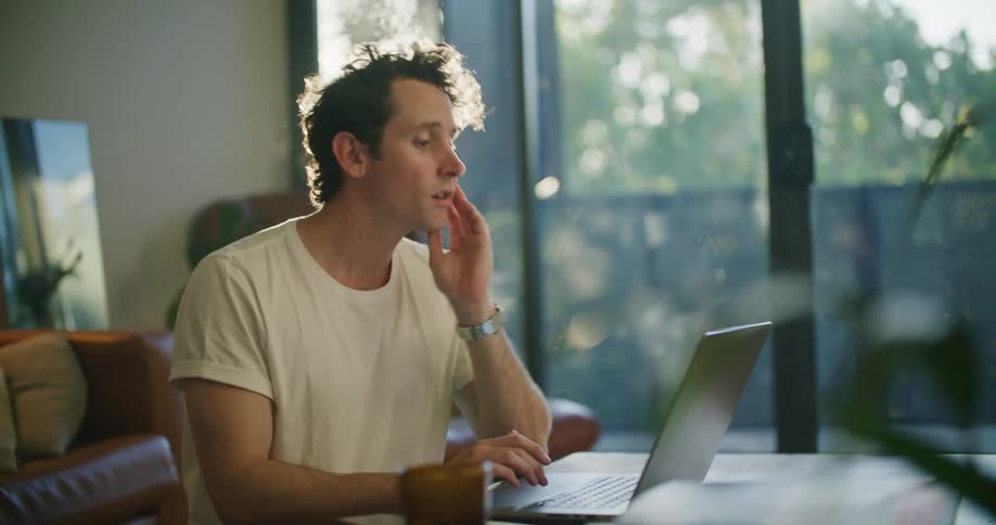 Man sitting at the table next to a glass door typing on his laptop and smiling