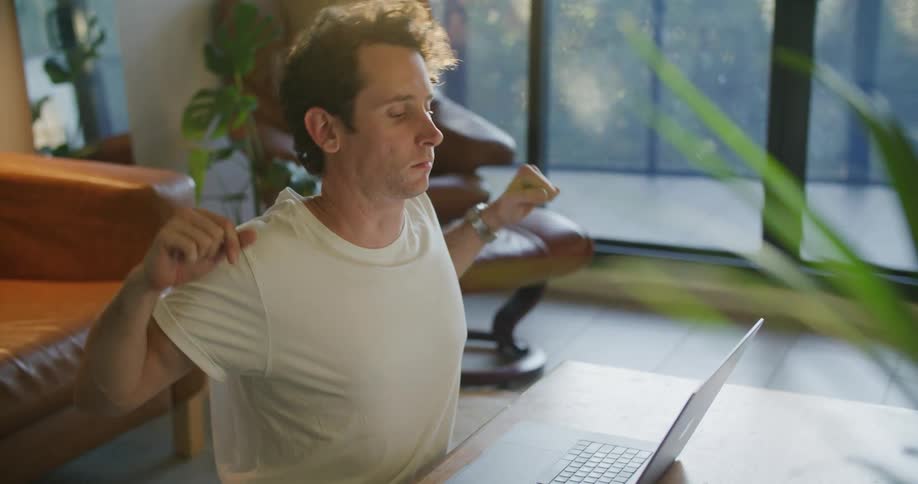 Young tired man stretches before continuing working on a laptop over a coffee table in the living room