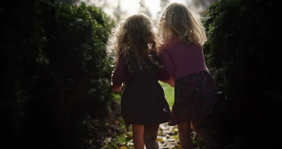 Two young blonde girls run towards the light through a secret garden holding hands