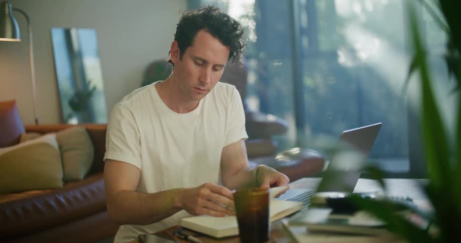 Man working from home opens a notebook in front of a laptop over the coffee table at morning