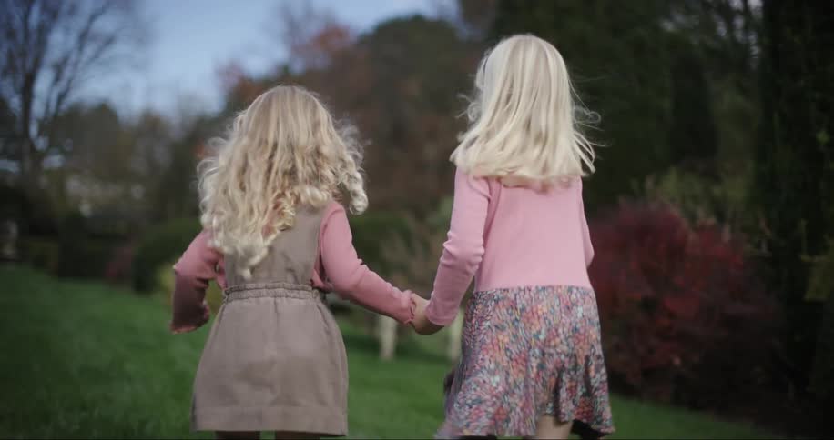 Two little blonde girls run through a garden holding hands with fall color