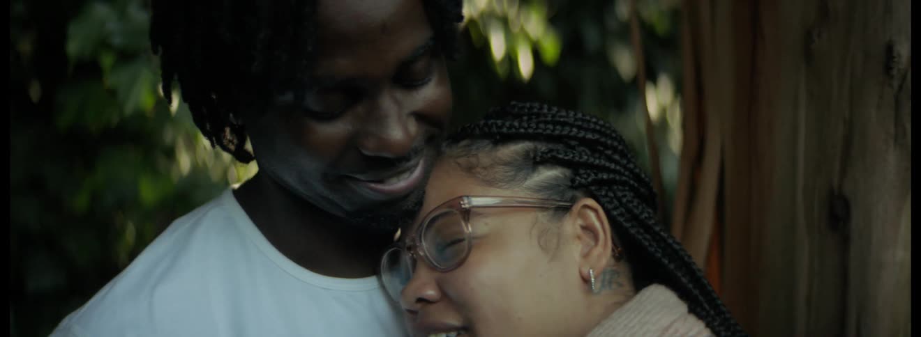 Man with dreadlocks and white t shirt holds and touches pregnant womans belly with glasses tattoo and earrings next to tree trunk
