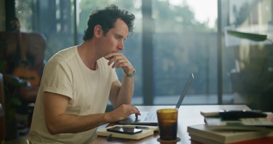 Man sitting at the table drinking from a cup typing on his laptop and writing down on a notepad