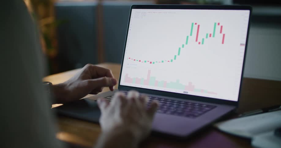 A man is sitting at a desk using a laptop computer with a graph on the screen