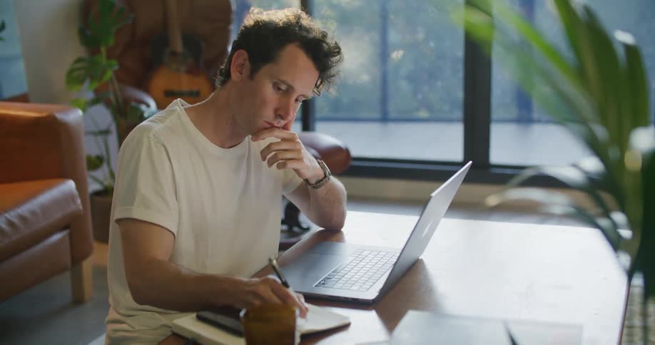 Man with white t shirt works on laptop and writing in notebookat coffee table next to brown couch in living room at home