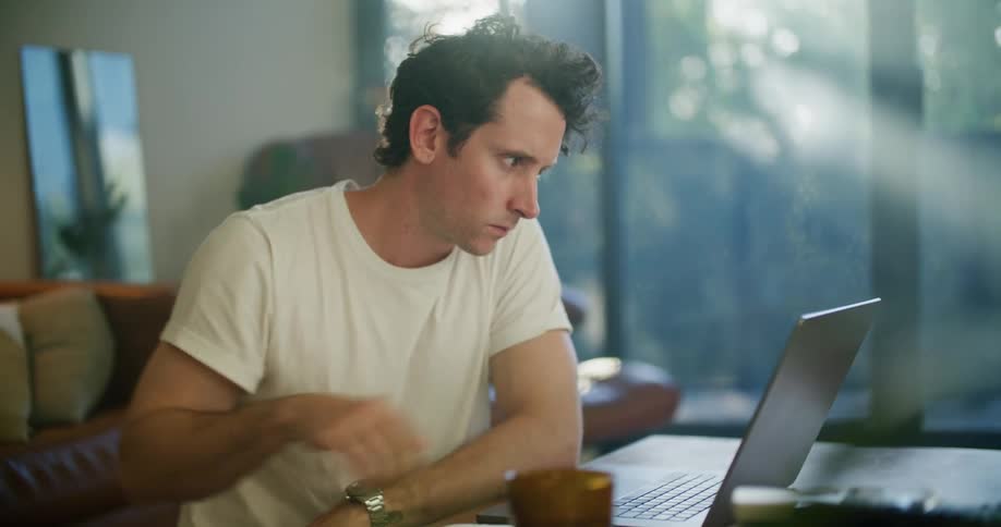 A man is sitting in his living room at a table working on a laptop computer