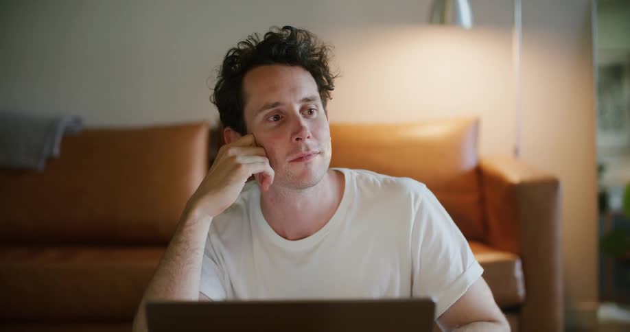 Man with white t shirt uses laptop in front of brown couch in living room at home