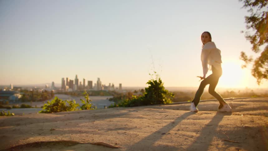 White woman dancing outdoors with the view of a big city in the background during sunset