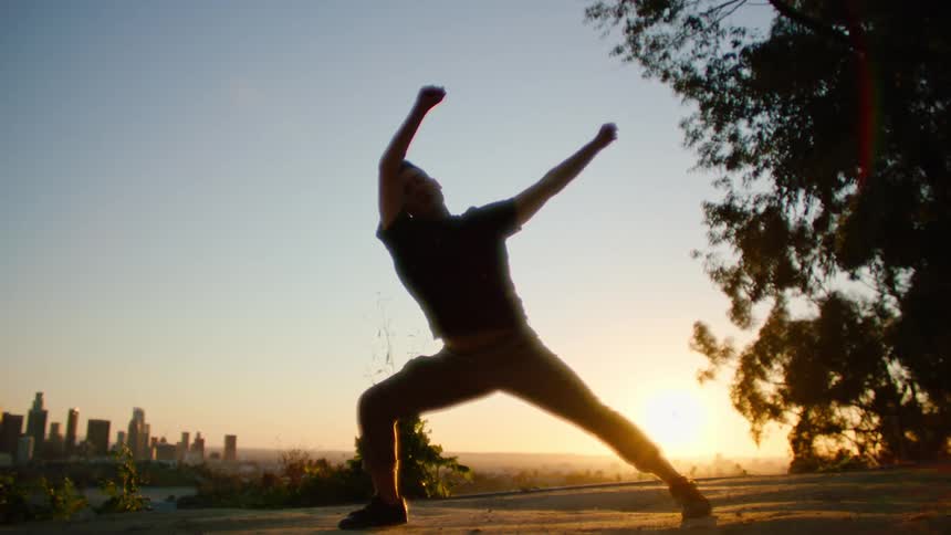 Dancing man wearing black t shirt and brown trousers is silhouetted against the city skyline and sunset