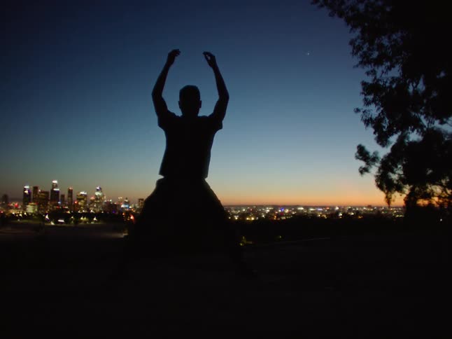Dancing man is silhouetted against the illuminated skyscrapers and sky at dusk