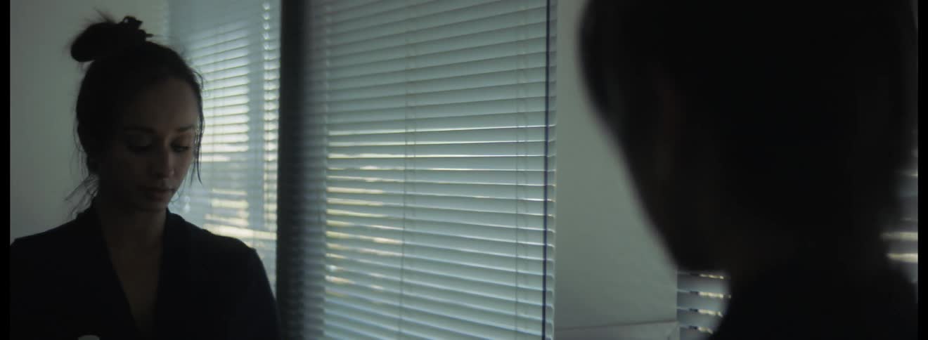 Woman with bun hair and black jacket looks in mirror by windows and blinds in the bathroom