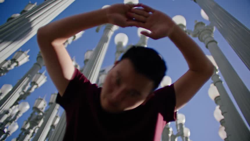 An asian male looks directly at the camera as he performs dance moves beneath many lamp posts