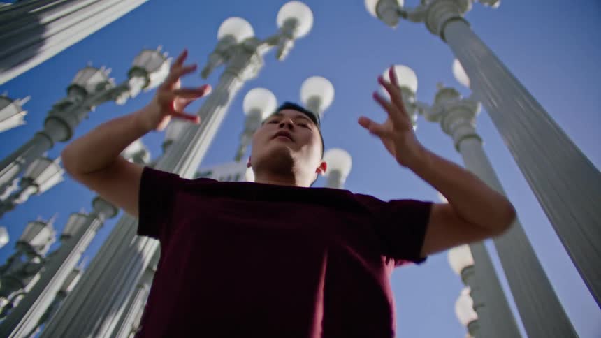 An asian male looks downward at the camera as he performs dance moves while outdoors