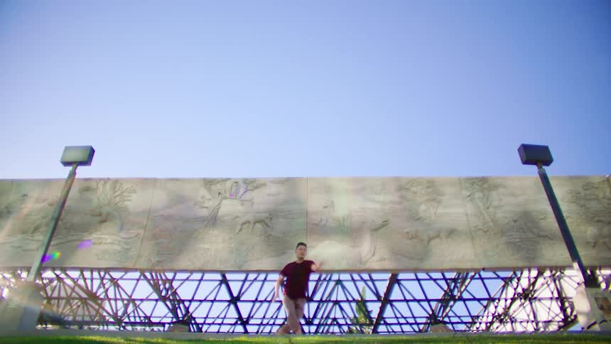 Man with red t shirt and brown pants dances in front of monument with animal and nature motifs