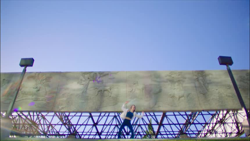 Female dancer dances near long panels of stone relief supported by metal struts under a clear blue sky