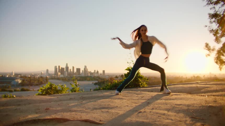 Female dancer dances outdoors back lit by sun sitting on the horizon with water and city skyline in the background