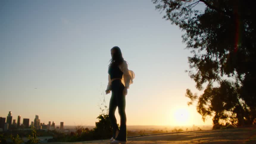 Dancing woman with white shirt black top and pants is silhouetted against the sunset and city skyline
