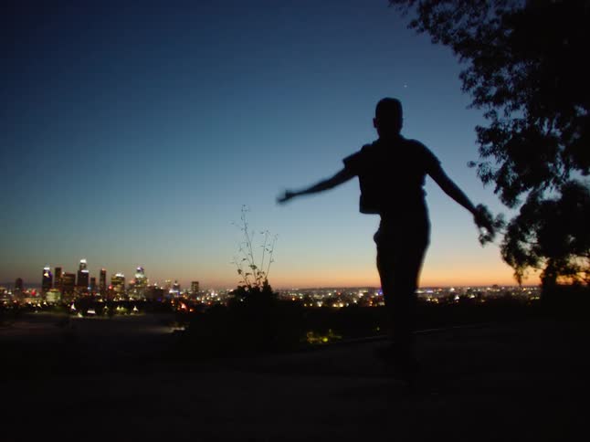 Male dancer dances as he is silhouetted against afterglow sitting on horizon behind city in the background