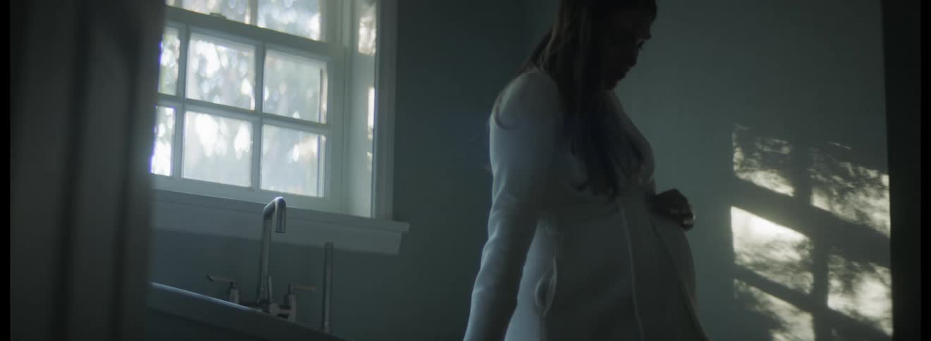 Pregnant woman with dark hair and white dress caresses her belly in the kitchen