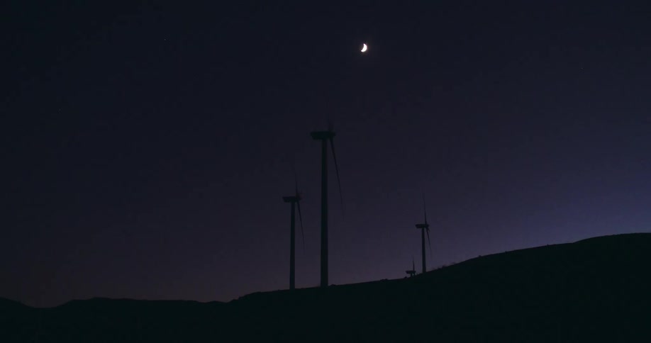 Wind turbines are silhouetted against the sky and moon at night on wind farm