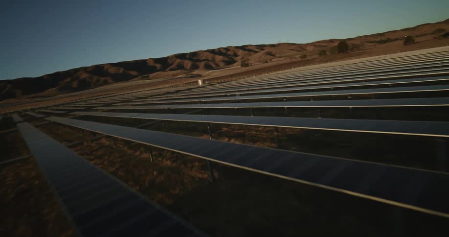 The sun shines brightly on solar panels on a solar farm with mountains in the distance