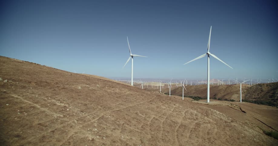 Wind turbines are spinning on a wind farm along hills and mountains in the distance under a clear sky