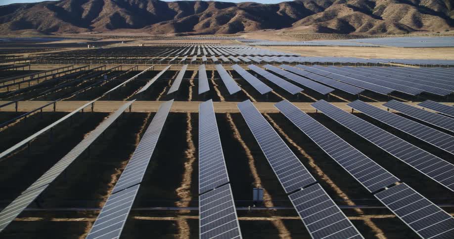 The sun shines brightly on solar panels on a solar farm surrounded by mountains